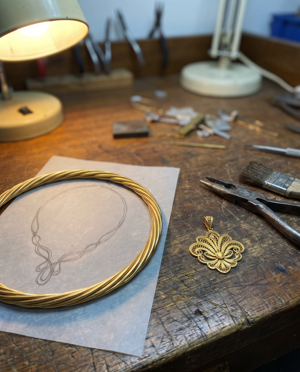 A pencil drawing of a necklace on tracing paper beside the finished gold piece on a workbench